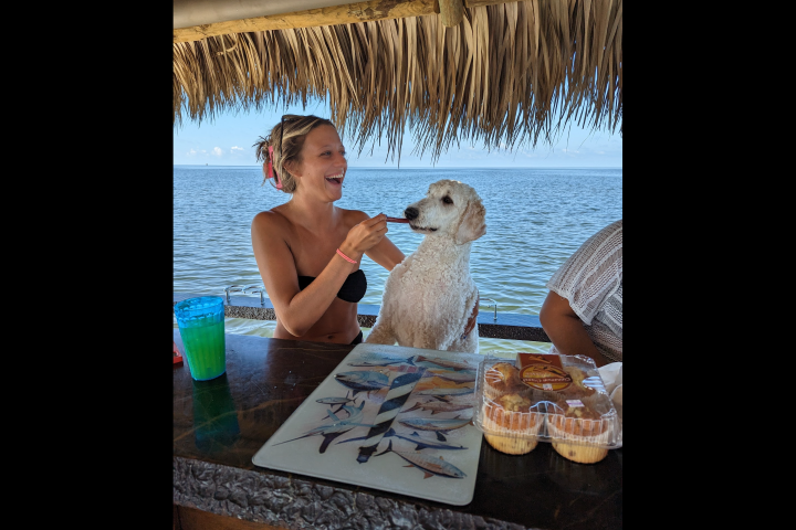 Woman in bikini and dog at beachside bar, colorful drink and food on counter.
