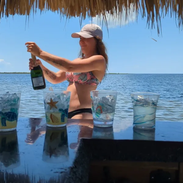 Woman in swimsuit opening champagne near water, tiki hut overhead, glasses on bar.