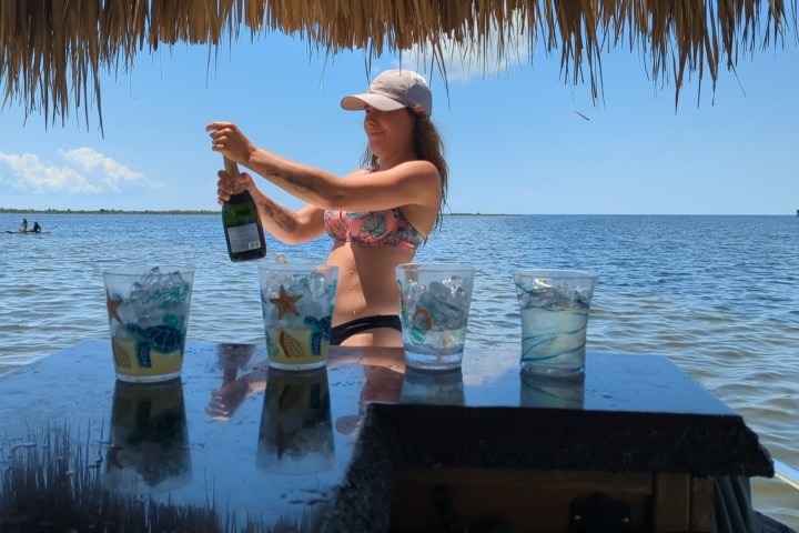 Woman in swimsuit opening champagne near water, tiki hut overhead, glasses on bar.