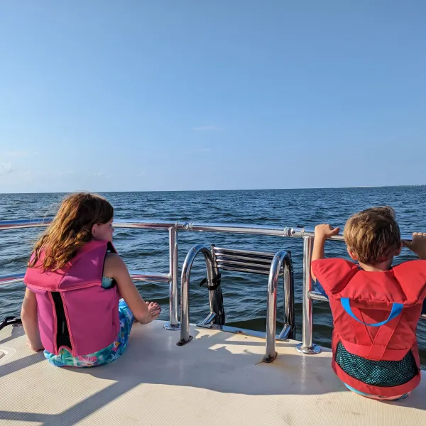 Two children in life jackets sitting on a boat, looking at the ocean.