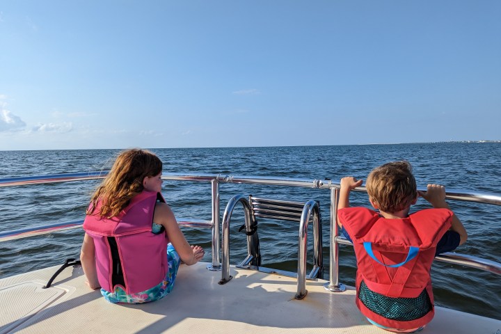 Two children in life jackets sitting on a boat, looking at the ocean.