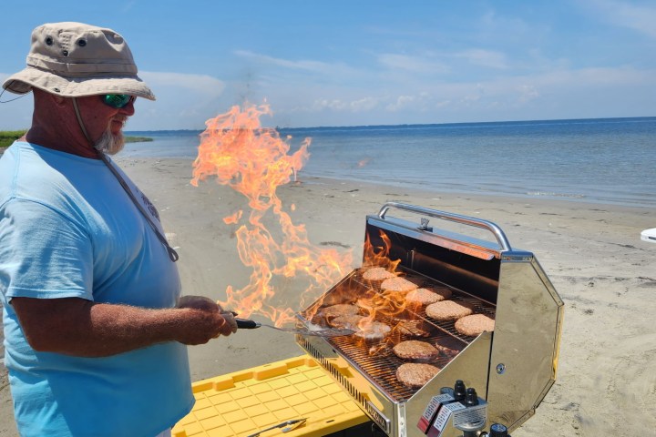 Man grilling burgers on a beach with flames rising from a barbecue.