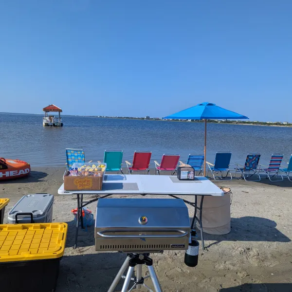 Beach chairs and umbrella by the water with a grill and inflatable raft on the sand.