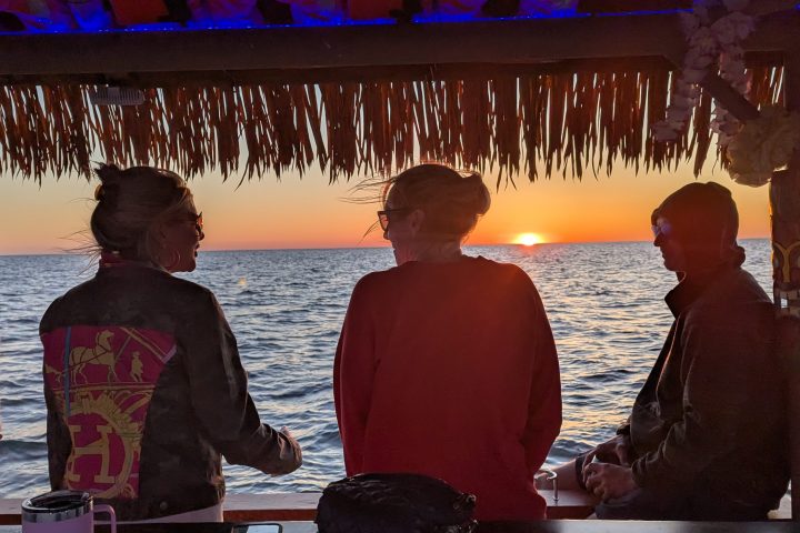 People under thatched roof enjoying sunset over ocean, seen from a bar or deck.