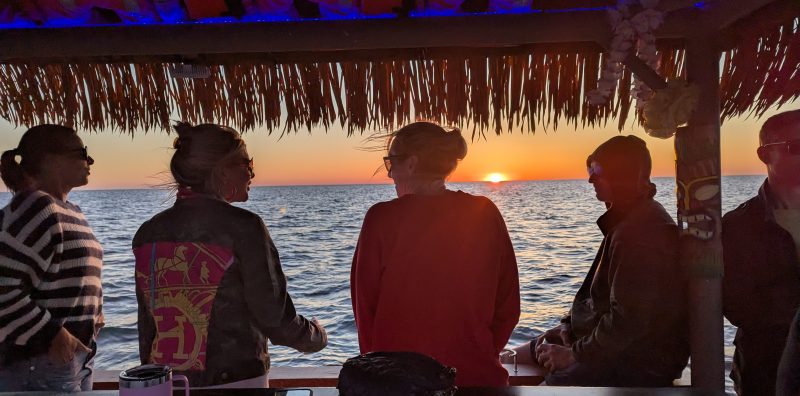 People under thatched roof enjoying sunset over ocean, seen from a bar or deck.
