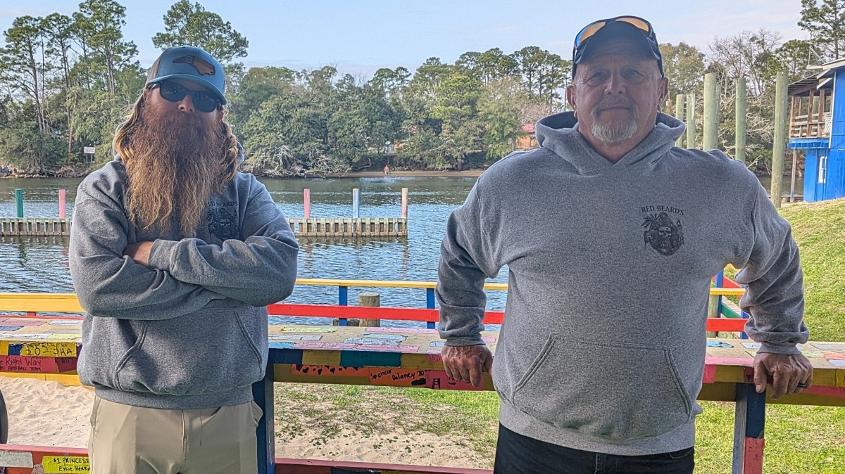 Two men in hoodies stand by a colorful railing with a river and trees in the background.