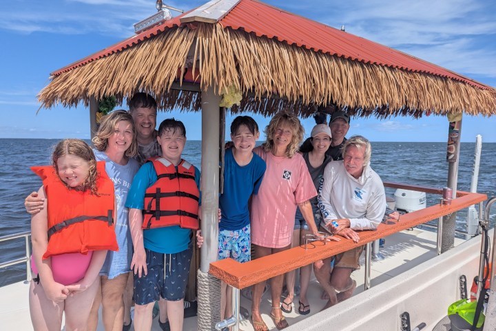 Group of people smiling on a boat with a thatched roof, wearing casual summer clothes.