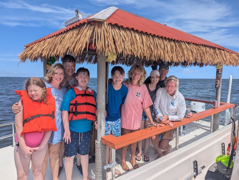 Group of people smiling on a boat with a thatched roof, wearing casual summer clothes.