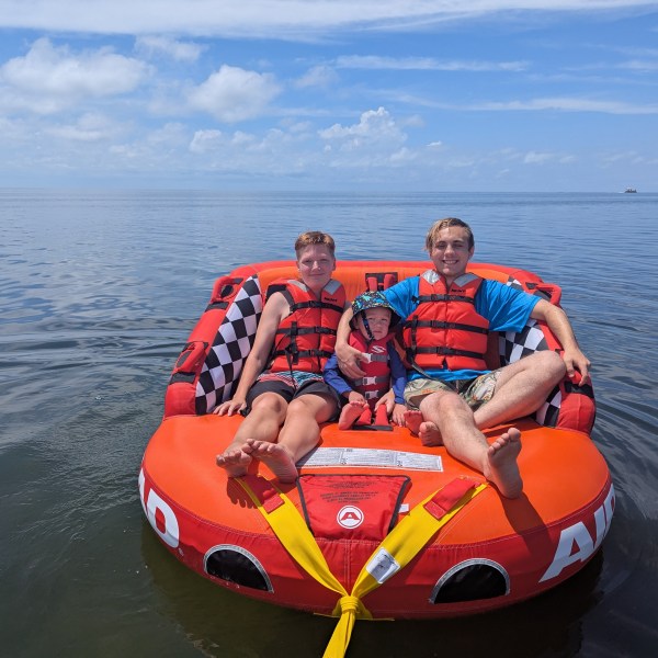 Three people wearing life jackets sitting on an inflatable raft on a calm, open sea under a blue sky.