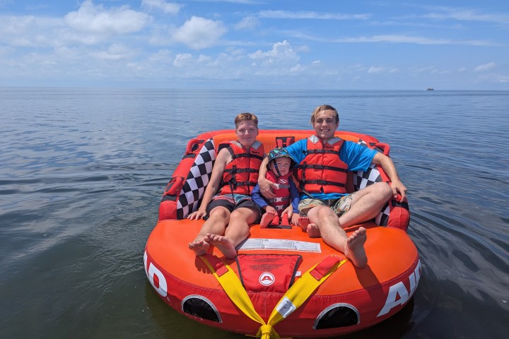 Three people wearing life jackets sitting on an inflatable raft on a calm, open sea under a blue sky.