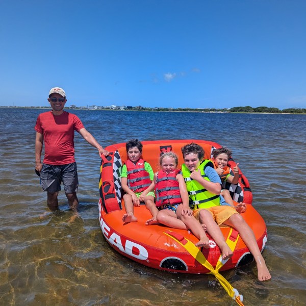 Smiling adult and four children on an inflatable raft in shallow water under a clear blue sky.
