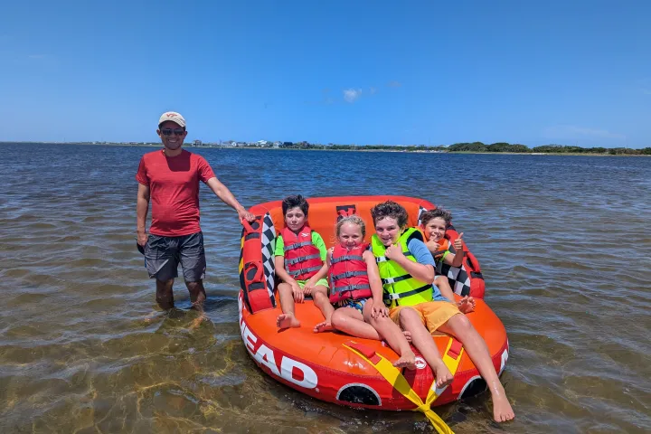 Smiling adult and four children on an inflatable raft in shallow water under a clear blue sky.