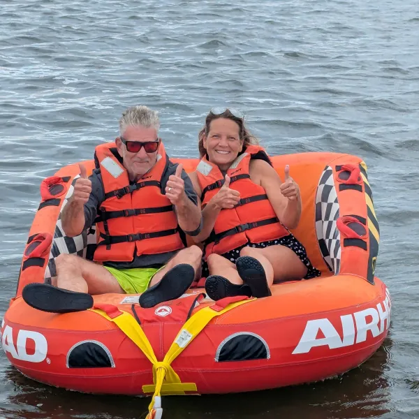 Two people in life jackets give thumbs up while sitting on an inflatable tube in water.