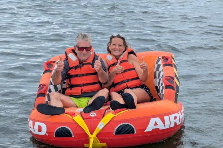 Two people in life jackets give thumbs up while sitting on an inflatable tube in water.
