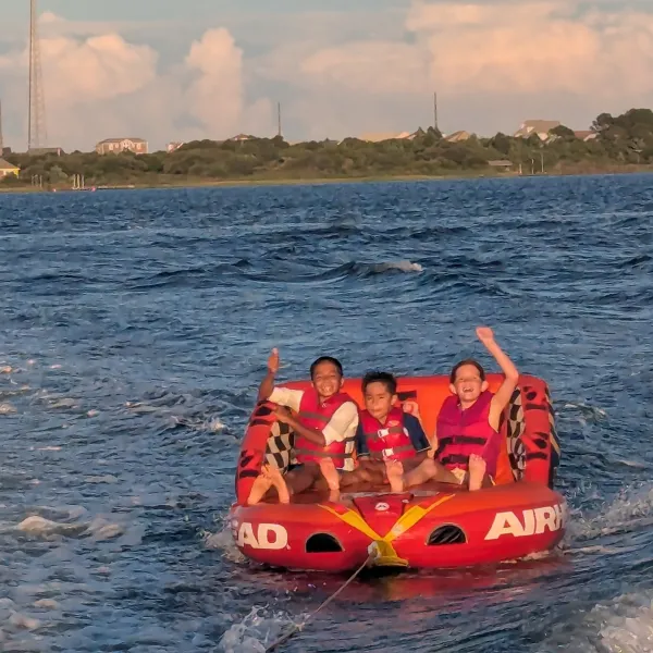 Three children in life vests riding a red inflatable tube on a lake, waving and smiling.