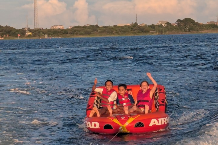 Three children in life vests riding a red inflatable tube on a lake, waving and smiling.