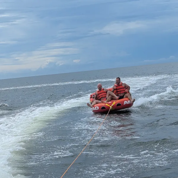 Two people tubing on water with orange life jackets, pulled by a boat under a cloudy sky.