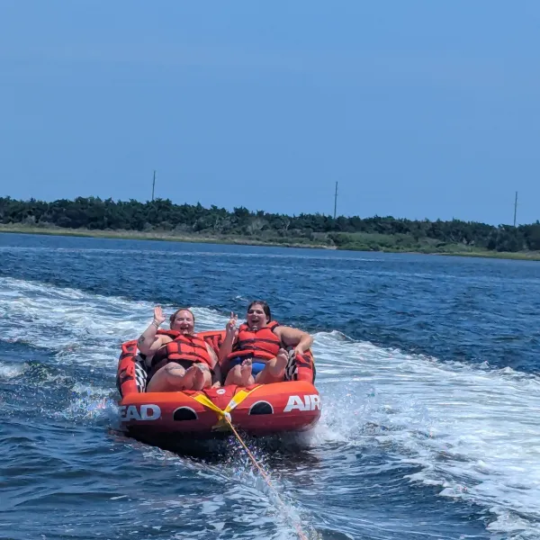 Two people in life vests ride an inflatable tube on water, smiling and waving.