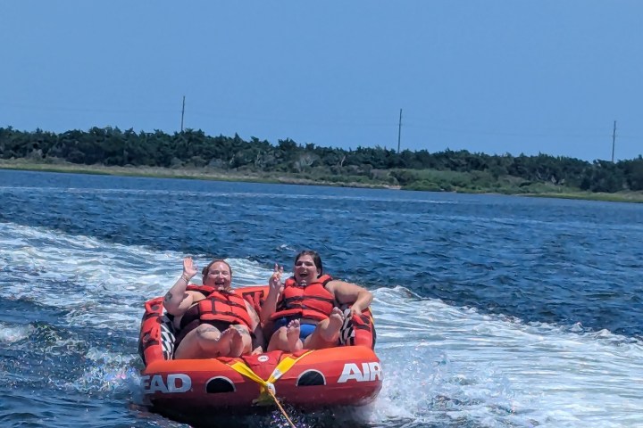 Two people in life vests ride an inflatable tube on water, smiling and waving.