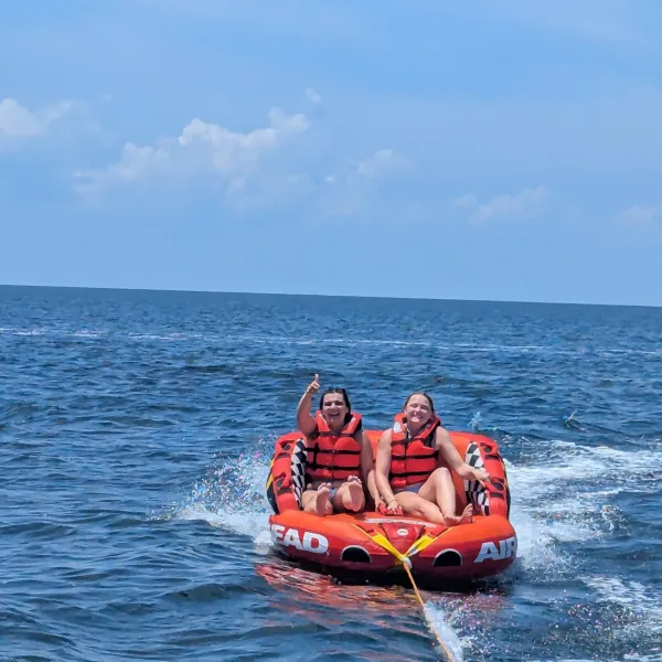 Two people in life vests on an inflatable tube being towed behind a boat on the ocean.
