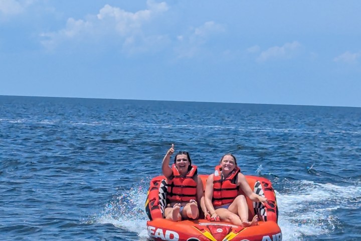 Two people in life vests on an inflatable tube being towed behind a boat on the ocean.