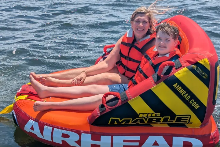 Two people smiling on an inflatable raft on the water, wearing orange life vests.