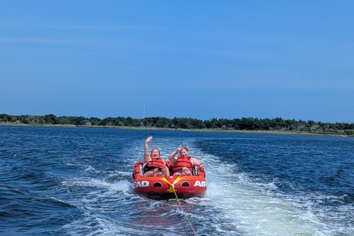 Two people tubing on a red inflatable, being pulled by a boat on a lake.