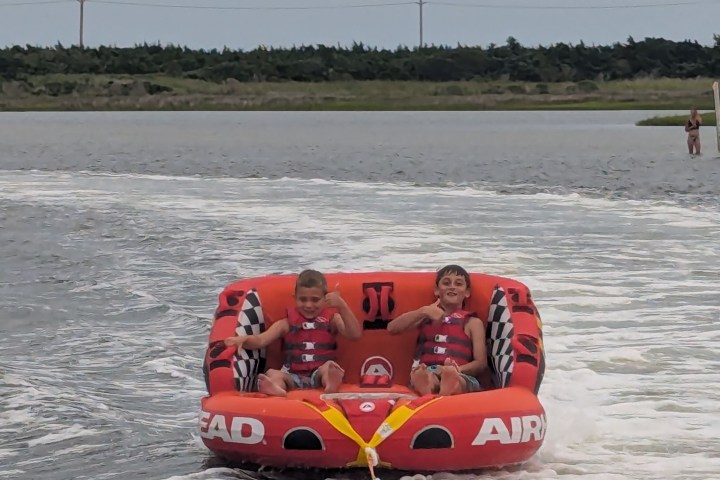 Two kids in red life vests on a red inflatable being towed on water.