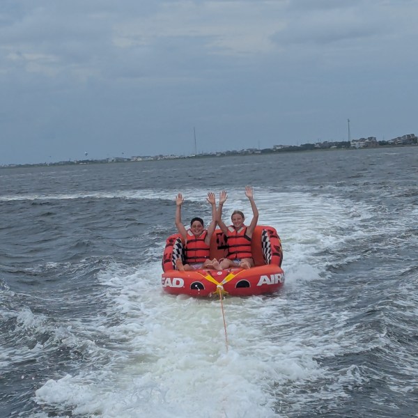 Two people on an inflatable tube being towed on water, both wearing life vests and raising their arms.