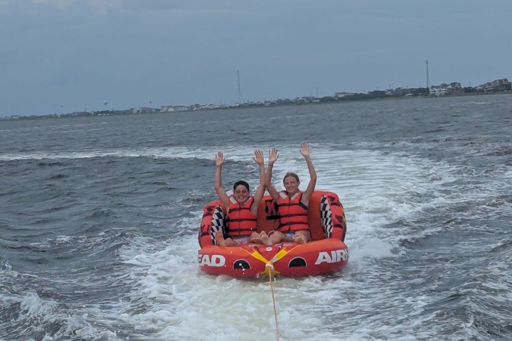 Two people on an inflatable tube being towed on water, both wearing life vests and raising their arms.