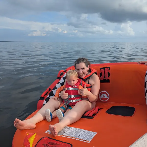 Person and child in life jackets sitting on an orange raft on a calm sea with cloudy sky.