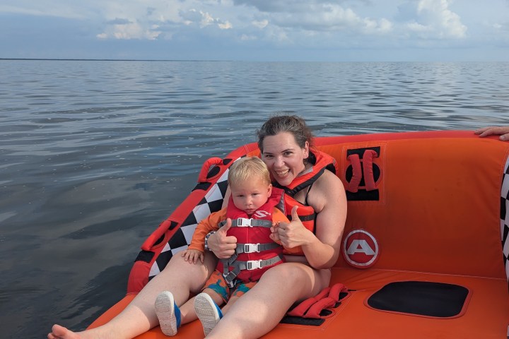 Person and child in life jackets sitting on an orange raft on a calm sea with cloudy sky.