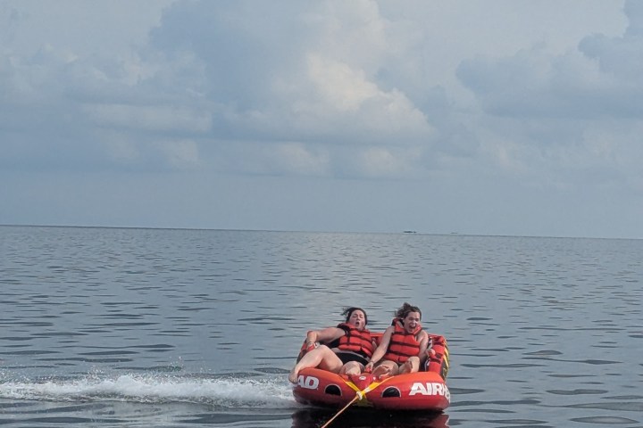 Two people in life vests tubing on water behind a boat under cloudy sky.
