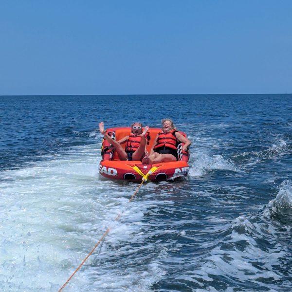 Two people tubing on ocean, wearing life jackets, laughing with waves behind them.