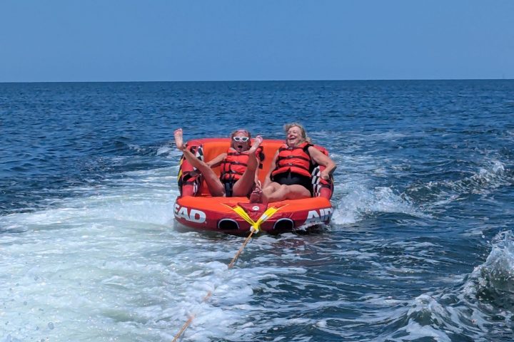 Two people tubing on ocean, wearing life jackets, laughing with waves behind them.
