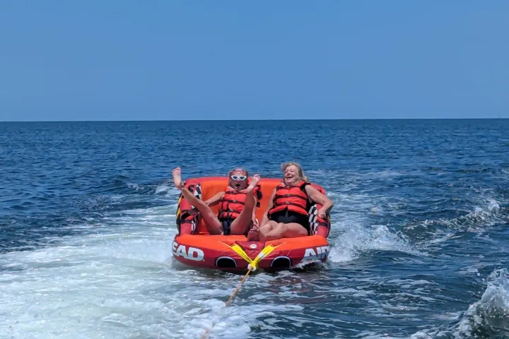 Two people tubing on ocean, wearing life jackets, laughing with waves behind them.