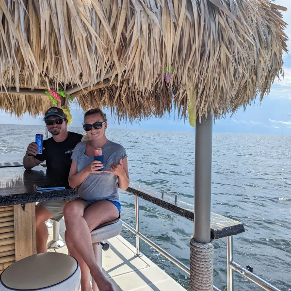 Two people sitting at a tiki bar on a boat, holding drinks, with ocean in the background.