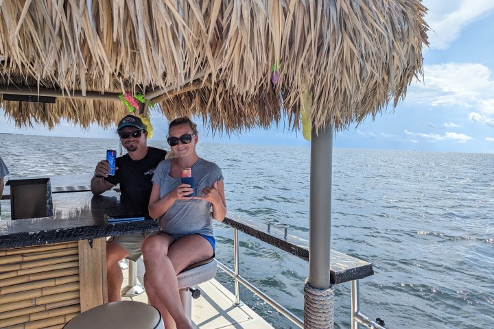 Two people sitting at a tiki bar on a boat, holding drinks, with ocean in the background.