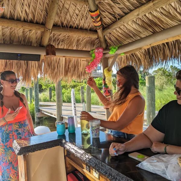 People at a tiki bar with drinks, surrounded by tropical decor under a thatched roof.