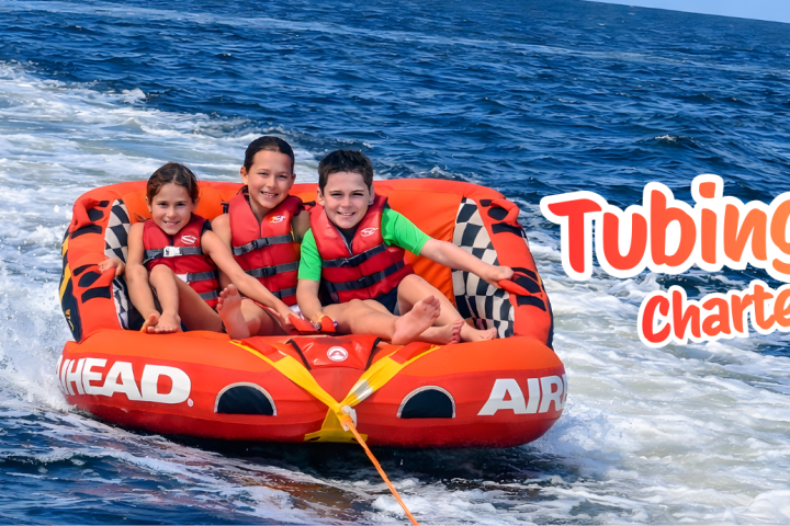 Three kids on a red inflatable tube being pulled on water with 'Tubing Charter' text on the side.
