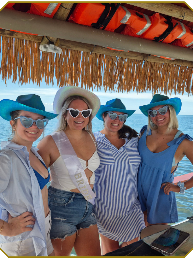 Four women in swimsuits and hats smiling at a beach bar under a thatched roof.