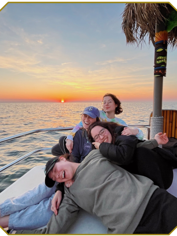 Four people laughing on a boat at sunset with a palm roof overhead.