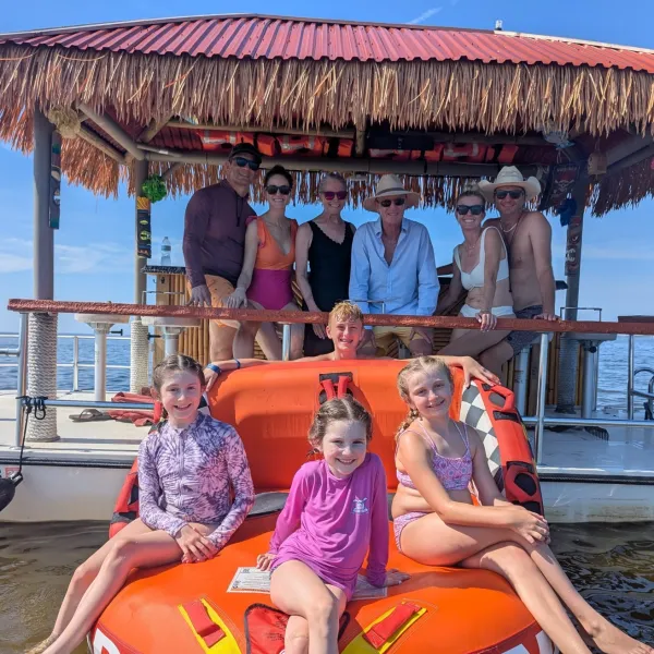 Group on a boat with a tiki roof over water, kids on an inflatable raft.