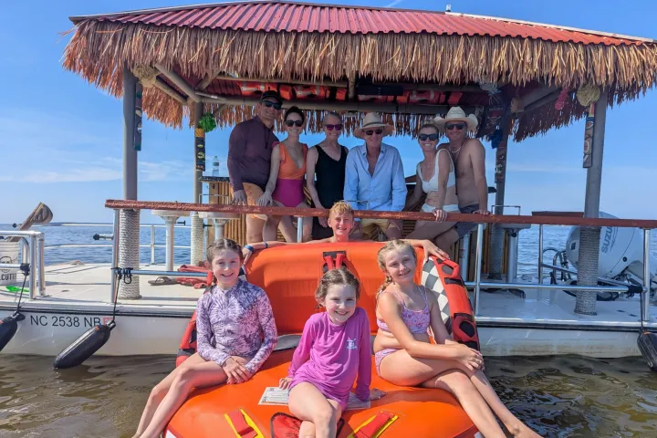 Group on a boat with a tiki roof over water, kids on an inflatable raft.