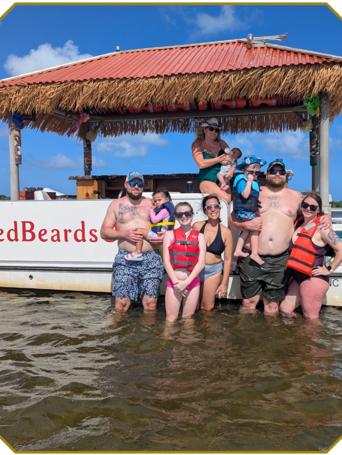 Group of people standing in shallow water by a boat with a thatched roof.