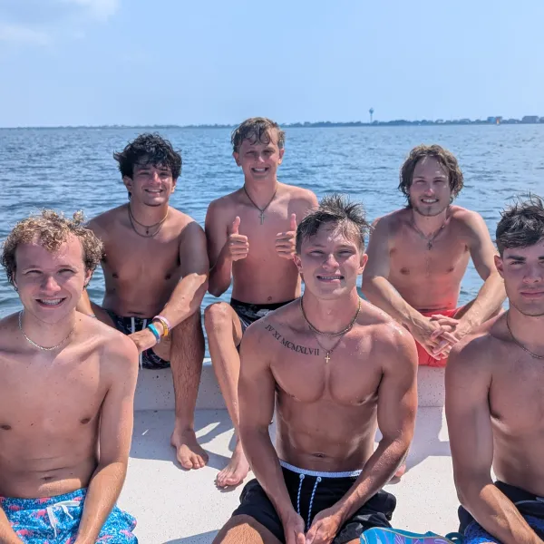 Six young men sitting shirtless on a boat with water and sky in the background.