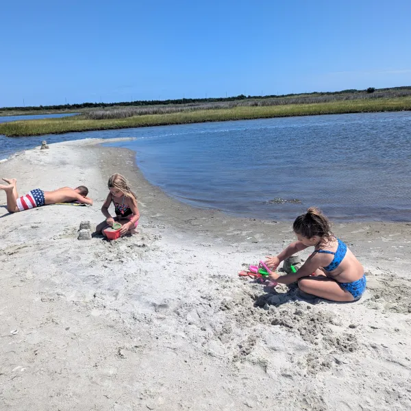 Three children play on a sandy beach by calm water under a clear blue sky.
