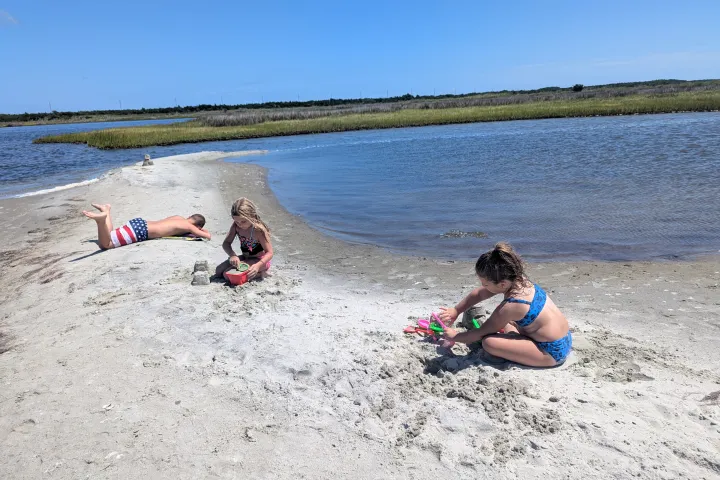 Three children play on a sandy beach by calm water under a clear blue sky.