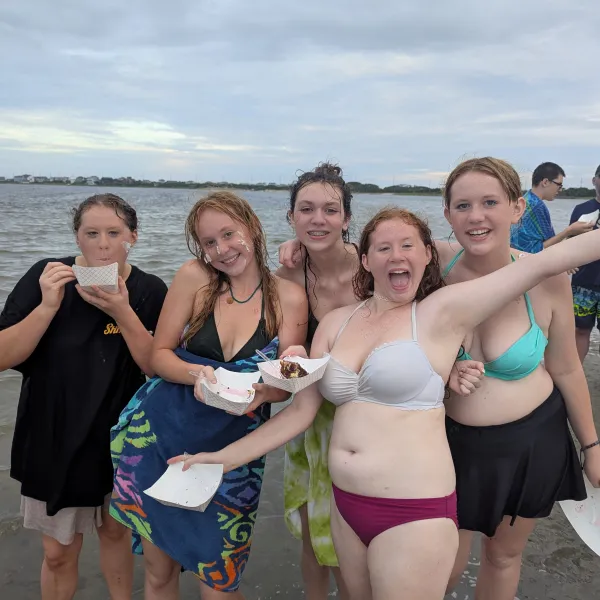 Five smiling people on a beach enjoying food, with a cloudy sky and water in the background.