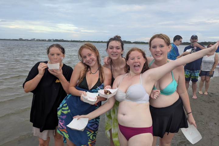 Five smiling people on a beach enjoying food, with a cloudy sky and water in the background.